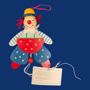 May include: A wooden clown ornament with a yellow hat, red hair, and a red nose. The clown has blue and white striped arms, blue pants with yellow stars, and a red belly with yellow stars. A tag is attached.