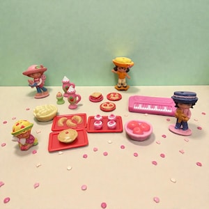 May include: A collection of miniature plastic toys featuring figures and food items. The toys include figures wearing hats, a pink piano, trays of pastries, and small desserts. The color palette is primarily pink, red, yellow, and green.