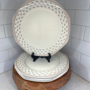 Może przedstawiać: Stos białych ceramicznych talerzy obiadowych z delikatnym wzorem kwiatowym w odcieniach niebieskiego i brązowego. Talerze są ułożone na drewnianym stojaku z trzema nogami.