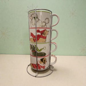 May include: A set of four white ceramic mugs stacked in a metal rack. Each mug features a different dog breed, adorned in festive winter attire. The dogs include a poodle, a golden retriever, a schnauzer, and a bulldog. The mugs are designed with a holiday theme.
