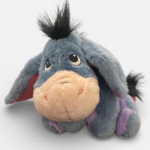May include: A plush Eeyore toy in a lying position. The donkey has blue fur, a pink muzzle, and a black tuft of hair. Purple stripes accent the legs. The toy has large, sad eyes and a small black tail.