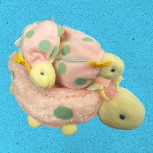 May include: A plush turtle-shaped pouch in soft pink with light green polka dots and a pale yellow head, feet, and tail. The shell features a zipper closure with yellow cord pulls, and the fabric has a textured, fuzzy finish throughout.