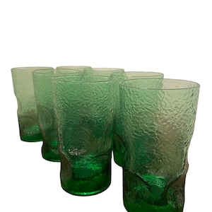 Op de afbeelding: Set van acht groene glazen tumblers met een getextureerd oppervlak. De glazen zijn in een rij gerangschikt, met de bodems van de glazen zichtbaar.