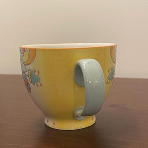 May include: A yellow ceramic mug with a light blue handle. The mug features a floral design in orange, blue, and red. The rim is outlined in orange, and the base has pink polka dots. The mug is on a brown surface.