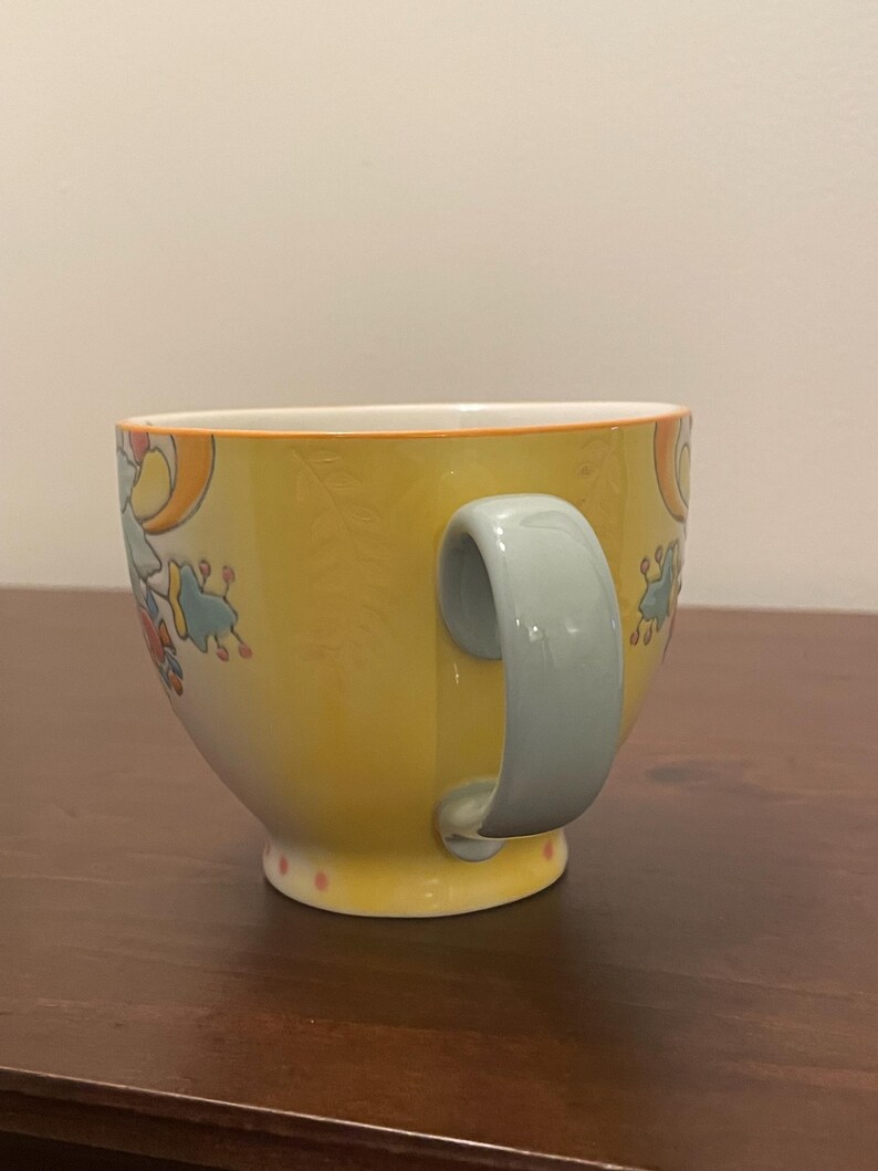 May include: A yellow ceramic mug with a light blue handle. The mug features a floral design in orange, blue, and red. The rim is outlined in orange, and the base has pink polka dots. The mug is on a brown surface.