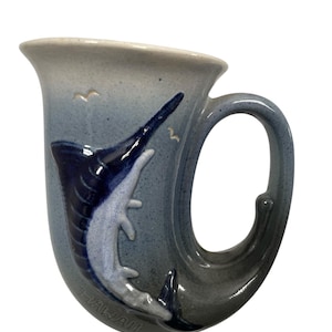 May include: A blue and white ceramic mug shaped like a marlin fish. The mug has a handle that is part of the fish's tail. The mug has the word "HAWAII" written on the bottom.