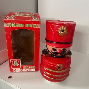 Könnte beinhalten: Eine rot-goldene Soldaten-Dekoration mit schwarzem Schnurrbart und roter Mütze. Der Soldat steht neben einer roten Schachtel mit den Worten "REVOLVING MUSICAL" und "SOLDIER DECORATION".