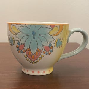 May include: A large ceramic mug with a floral design. The mug features a light blue, orange, and yellow floral pattern on a white background. The handle and rim are light blue and the base has small pink dots.