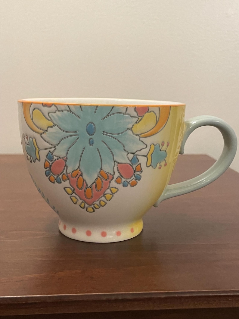 May include: A large ceramic mug with a floral design. The mug features a light blue, orange, and yellow floral pattern on a white background. The handle and rim are light blue and the base has small pink dots.