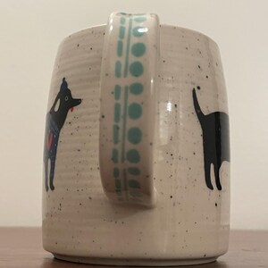 May include: A speckled ceramic mug with a teal handle and dog illustrations. One dog is black with a blue and red design, the other is a black silhouette. The handle has a teal geometric pattern.