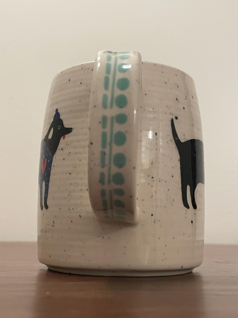 May include: A speckled ceramic mug with a teal handle and dog illustrations. One dog is black with a blue and red design, the other is a black silhouette. The handle has a teal geometric pattern.