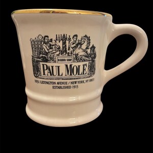 May include: White ceramic mug with a gold rim and a black and white illustration of a barbershop scene. The mug has the text "BARBER SHOP PAUL MOLE 1031 LEXINGTON AVENUE/NEW YORK, NY 10021 ESTABLISHED 1913" printed on it.