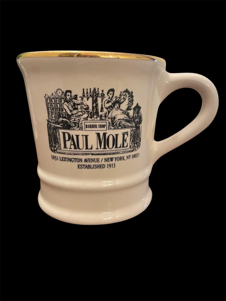 May include: White ceramic mug with a gold rim and a black and white illustration of a barbershop scene. The mug has the text "BARBER SHOP PAUL MOLE 1031 LEXINGTON AVENUE/NEW YORK, NY 10021 ESTABLISHED 1913" printed on it.