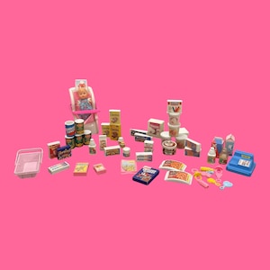 Pode incluir: Uma coleção de mantimentos em miniatura, incluindo caixas de cereais, conservas e produtos lácteos, dispostos em uma superfície rosa. Uma boneca está sentada em uma cadeira alta, e uma caixa registradora azul também está presente. Vários brinquedos de comida estão espalhados.