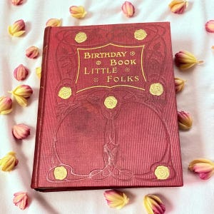 May include: A vintage red book titled "Birthday Book Little Folks" with gold lettering and decorative accents. The book is surrounded by small pink and yellow flower buds, creating a soft, romantic aesthetic.