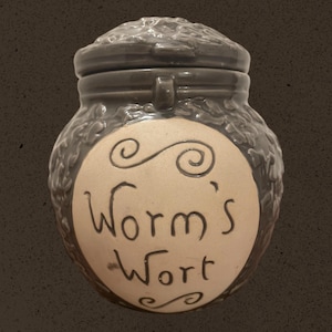 May include: A dark gray ceramic jar with a lid, featuring the text "Worm's Wort" on a cream-colored circular label. The jar has a textured, organic design and a small handle on the front.