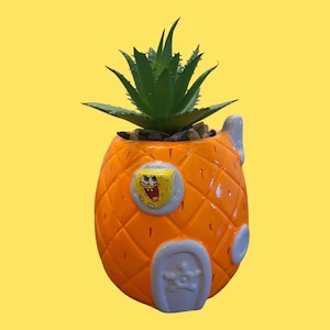 May include: An orange pineapple-shaped ceramic planter featuring a raised yellow Spongebob character face, a light purple door, and a light purple window. The planter contains a green succulent plant with pointed leaves and small brown pebbles.