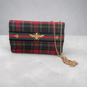 May include: A rectangular clutch bag in a red and black tartan pattern. It has a black velvet stripe with red and green stripes, decorated with gold-coloured bee motifs. A gold chain strap is attached.