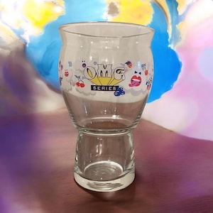 May include: A clear glass with a wide top and a smaller base. The glass features the text "OMG SERIES" and cartoon graphics of cherries, lips, and other designs. The glass sits on a purple surface with a colourful background.