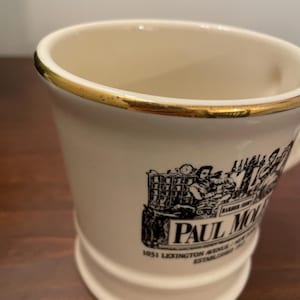 May include: White ceramic mug with a gold rim and a black and white illustration of a man working at a counter. The mug has the text "Paul Molitor 1051 Lexington Avenue, New York, NY Established 1903" printed on the side.