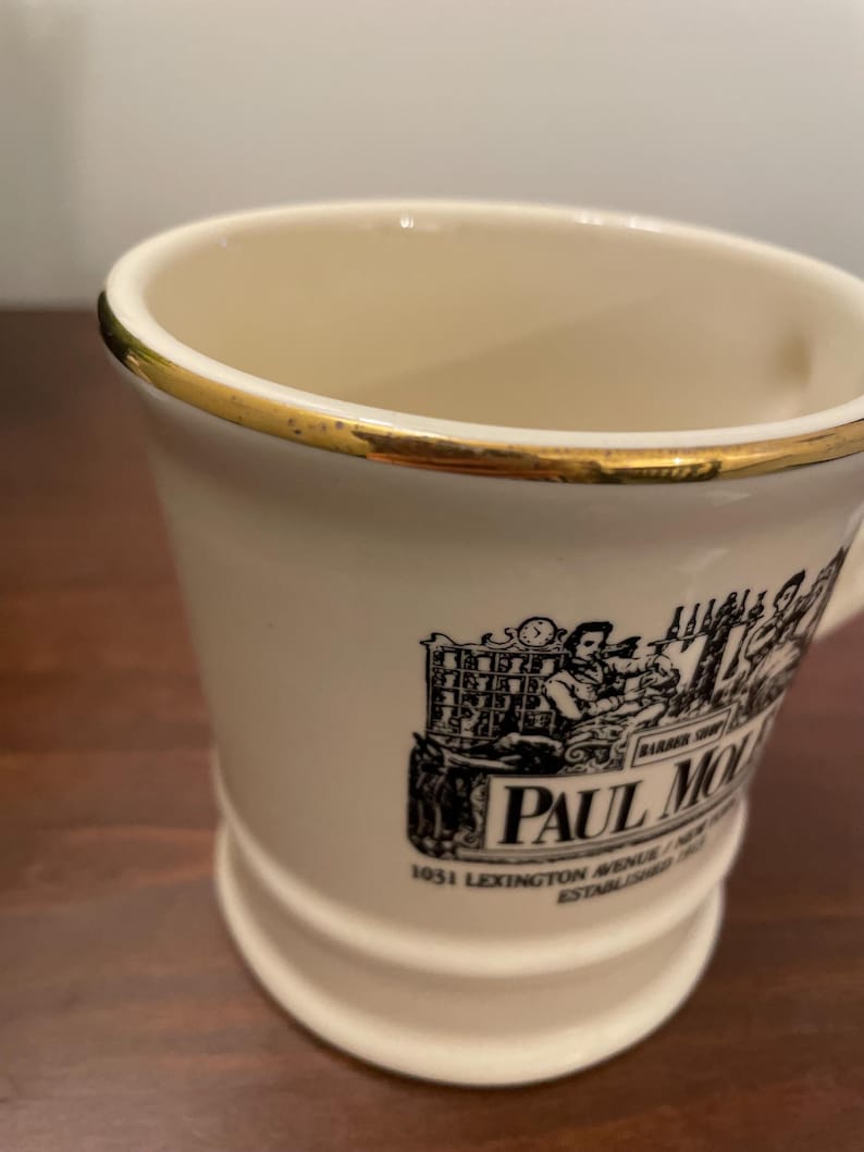 May include: White ceramic mug with a gold rim and a black and white illustration of a man working at a counter. The mug has the text "Paul Molitor 1051 Lexington Avenue, New York, NY Established 1903" printed on the side.