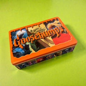 May include: An orange rectangular tin box with the words "Goosebumps" in an orange, stylized font. The box features illustrations of monsters and scenes from the book series. The box is on a green surface.