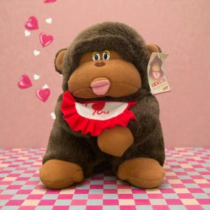 Op de afbeelding: Een pluche gorilla speelgoed met bruine vacht, een bruin gezicht en een rode slabbetje. De slabbetje heeft een wit midden met de woorden "I Love You" erop gedrukt. Het speelgoed heeft een label met de tekst "SENSOR Whistle Monkey".