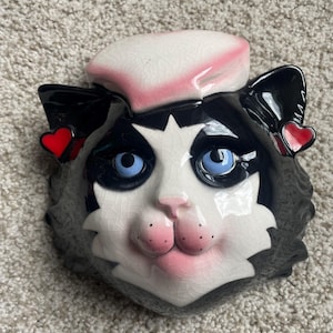 Könnte beinhalten: Keramik-Katzenkopf mit rosa Barett und roten Herzakzenten. Die Katze hat schwarz-weißes Fell, blaue Augen und rosa Lippen und Nase. Das Katzengesicht ist ein dekorativer Gegenstand.