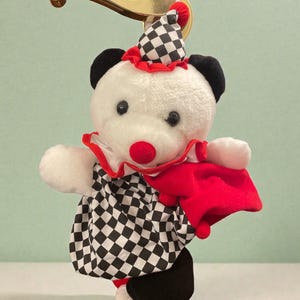 May include: A plush clown bear toy with a white body, black ears, and a red nose. It wears a checkered black and white outfit, a red collar, and a matching hat. The bear is hanging from a gold-colored handle.