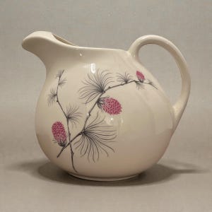 May include: Cream ceramic pitcher with a curved handle and spout. The pitcher is decorated with a black and grey branch design and pink pine cones. The pitcher has a glossy finish, likely for serving beverages.