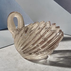 May include: A translucent, light brown glass swan figurine featuring a moulded, ribbed texture that mimics feathers along its body. The swan has a curved neck and a smooth, rounded base, with light reflecting off the faceted glass surfaces.