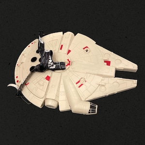 May include: A cream-colored Millennium Falcon shaped bag featuring molded mechanical details and red accents. A black lanyard with the white text Star Wars is attached to the top. The bag has a circular main body with a protruding cockpit section.