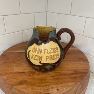 May include: A ceramic pitcher with a brown handle and a spout. The pitcher is primarily beige with brown and gray accents. The text "OHNE FLEISS KEIN PREIS" is displayed on the front of the pitcher. The pitcher is sitting on a wooden stand.