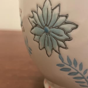May include: A white ceramic mug with a floral design. The mug features a large, light blue flower with a dark outline, and smaller blue floral accents. The base has pink polka dots.