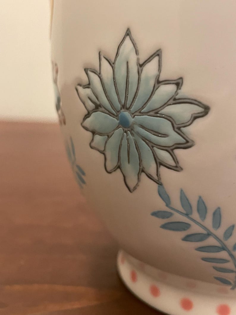 May include: A white ceramic mug with a floral design. The mug features a large, light blue flower with a dark outline, and smaller blue floral accents. The base has pink polka dots.