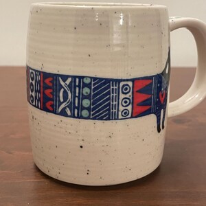 May include: A speckled ceramic mug with a handle. The mug features a band of blue, red, and white geometric patterns around its middle. The mug's exterior is a creamy white color with small black speckles.