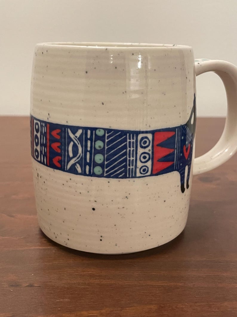 May include: A speckled ceramic mug with a handle. The mug features a band of blue, red, and white geometric patterns around its middle. The mug's exterior is a creamy white color with small black speckles.