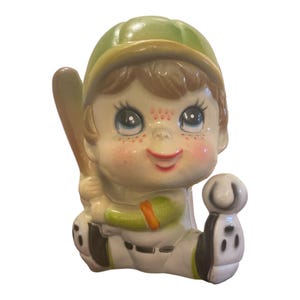 May include: A ceramic figurine of a baseball player wearing a green cap and white uniform with green accents. The figure has brown hair, freckles, and large eyes, holding a brown baseball bat in one hand and a white baseball in the other hand.