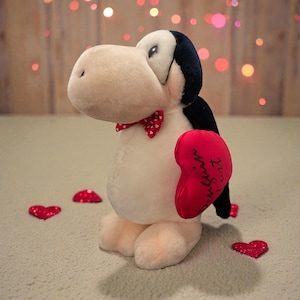 Pode incluir: Um brinquedo de pelúcia de pinguim com corpo creme, asas pretas e gravata borboleta vermelha. O pinguim segura uma almofada vermelha em forma de coração com texto em itálico. Há lantejoulas vermelhas em forma de coração espalhadas. O brinquedo tem cerca de 25 cm de altura.