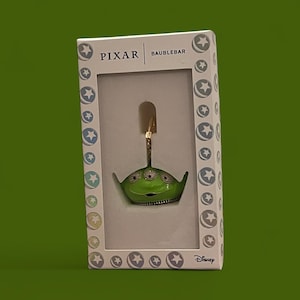 May include: A green alien-shaped ornament with three crystal eyes and a smiling mouth, presented in a decorative box. The box features the Pixar and Baublebar logos, along with star and circle patterns. The ornament has a gold-coloured loop for hanging.