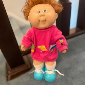 May include: A vintage Cabbage Patch Kid doll with fair skin, green eyes, and orange hair. The doll wears a pink sweatshirt with a small graphic and blue shoes with white laces. The doll is standing on a blue carpet.