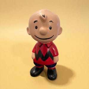 May include: A ceramic figurine of Charlie Brown from the Peanuts comic strip. The figure is wearing a red shirt with a black zig-zag pattern, blue shorts, and black shoes. The figurine has a smiling face and a small curl of hair.