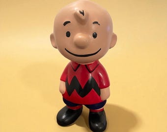 May include: A ceramic figurine of Charlie Brown from the Peanuts comic strip. The figure is wearing a red shirt with a black zig-zag pattern, blue shorts, and black shoes. The figurine has a smiling face and a small curl of hair.