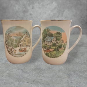 May include: Two white ceramic mugs with gold trim. The mugs feature images of a house in a winter scene and a house in a summer scene. The text on the mugs reads "Currier & Ives Winter" and "Currier & Ives Summer".