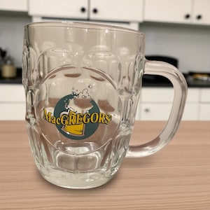 Puede incluir: Un vaso de cerveza transparente con asa. El vaso presenta un logotipo con el texto "MacGREGORS" en letras amarillas, un círculo verde y una ilustración de cerveza espumosa. El vaso está sobre una superficie marrón claro.