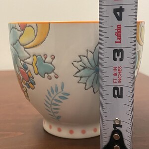 May include: White ceramic bowl with floral designs in blue, yellow, and orange. The bowl has an orange rim and a base with pink polka dots. The bowl is approximately 3 inches tall.
