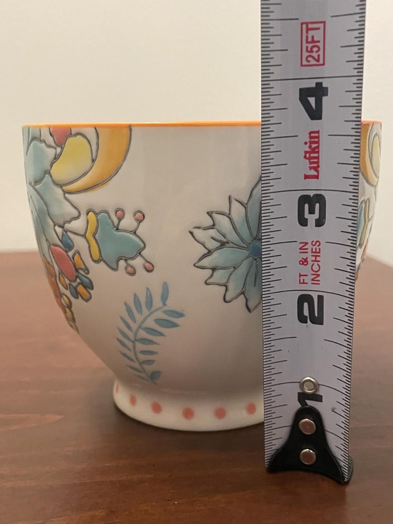 May include: White ceramic bowl with floral designs in blue, yellow, and orange. The bowl has an orange rim and a base with pink polka dots. The bowl is approximately 3 inches tall.