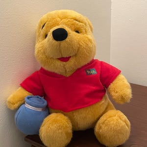 May include: A plush Winnie the Pooh toy, featuring a yellow body, black eyes, and a red shirt. The toy is holding a blue pot with a white ribbon. The Pooh logo is on the shirt.