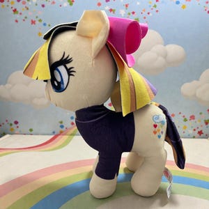May include: A plush toy pony with a cream-colored body, blue eyes, and a purple outfit. The pony has a yellow and rainbow mane and tail, and a pink bow. The tail features a cloud with colorful hearts.