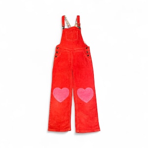 May include: Red corduroy overalls with pink heart patches on the knees. The overalls have adjustable straps with a floral pattern and button closures on the sides. A small pocket is on the chest.